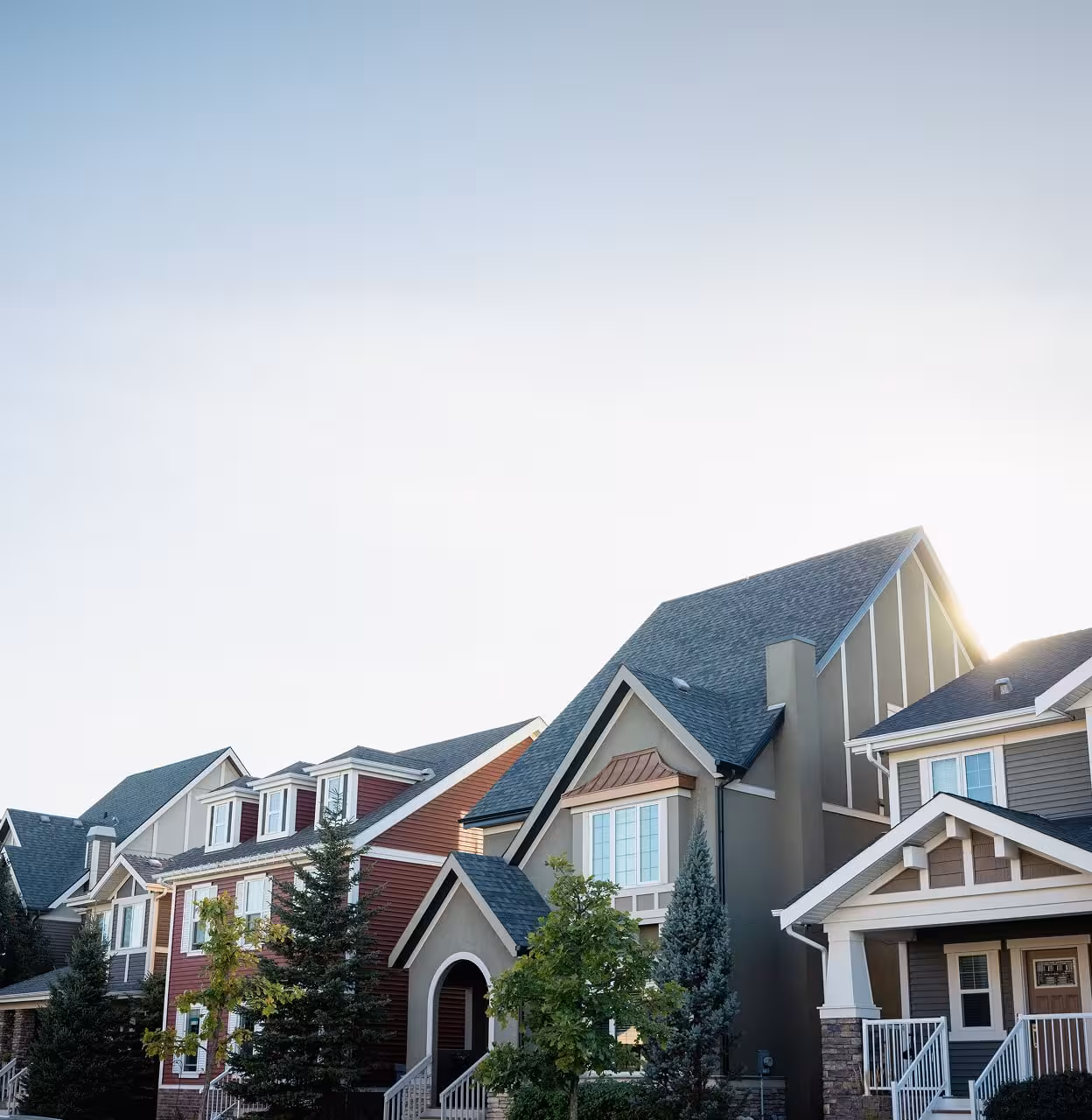 Row of colourful Griesbach homes with steep rooflines, front porches, trees, and a bright sky overhead.